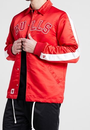 Homme portant une veste rouge Chicago Bulls avec des bandes blanches sur les manches et un pantalon noir, ajustant le devant de la veste.