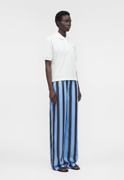 Γυναίκα όρθια, φοράει λευκό πόλο με κοντά μανίκια, φαρδιά μπλε παντελόνια με μαύρες κάθετες ρίγες και μαύρα σανδάλια, στραμμένη ελαφρά προς τα δεξιά.