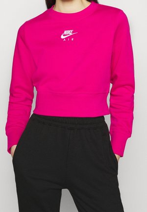 Osoba ubrana w jaskraworóżową krótka bluzę Nike Air i czarne spodnie, z rękami w kieszeniach, na tle jednolitego jasnego tła.