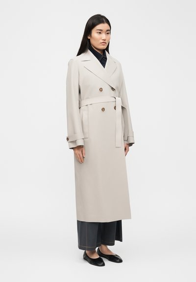 Γυναίκα με μακριά σκούρα μαλλιά φοράει μπεζ διπλόμπιστο τζάκετ τύπου trench πάνω από μαύρο παντελόνι φαρδύ και μαύρα φλατ παπούτσια, στέκεται μπροστά σε απλό φόντο.