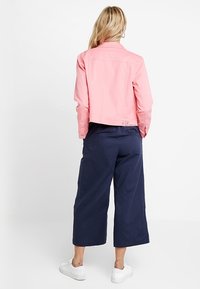 Chaqueta de mezclilla rosa recortada con cuello y detalles de botones, combinada con culottes de corte amplio en color navy y zapatillas blancas. Vista trasera.