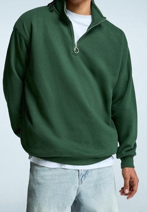 Personne portant un sweat-shirt à demi-fermeture éclair vert foncé sur une chemise blanche, associé à un jean bleu clair, debout devant un fond uni.
