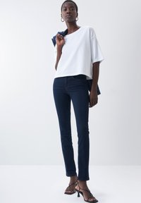 T-shirt blanc court en coton doux, associé à un jean ajusté bleu foncé. Le modèle porte des sandales à talons noirs à lanières et tient une veste en denim bleue.