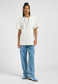 T-shirt oversize cotone color beige chiaro con maniche corte, scollatura rotonda e piccolo logo; abbinata a jeans baggy blu chiaro e sneakers blu.