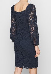 Navy blue lace dress with a square neck, long puff sleeves, and intricate floral patterns. Features a fitted waist and lace hem.