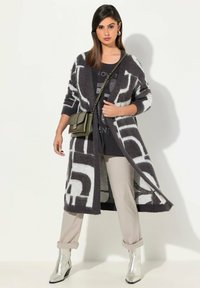 Langer, offener Cardigan in dunkelgrauem und weißem geometrischem Muster mit weicher Textur. Kombiniert mit hellen Hosen und glänzenden silbernen Ankle-Boots.