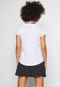 Žena s kudrnatými vlasy ve bílém sportovním tričku s krátkým rukávem a černé plisé sukni, stojící zády k fotografii na jednobarevném pozadí.