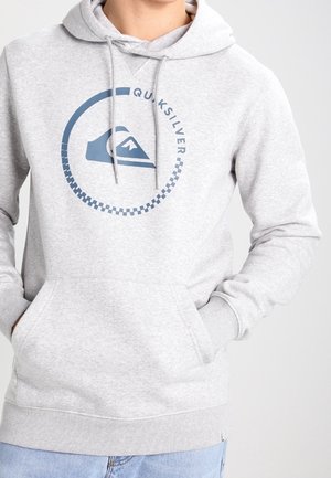 Persoon draagt een lichtgrijze Quiksilver-hoodie met een cirkelvormig logo en een blauwe spijkerbroek, handen in de voorzak, effen achtergrond.