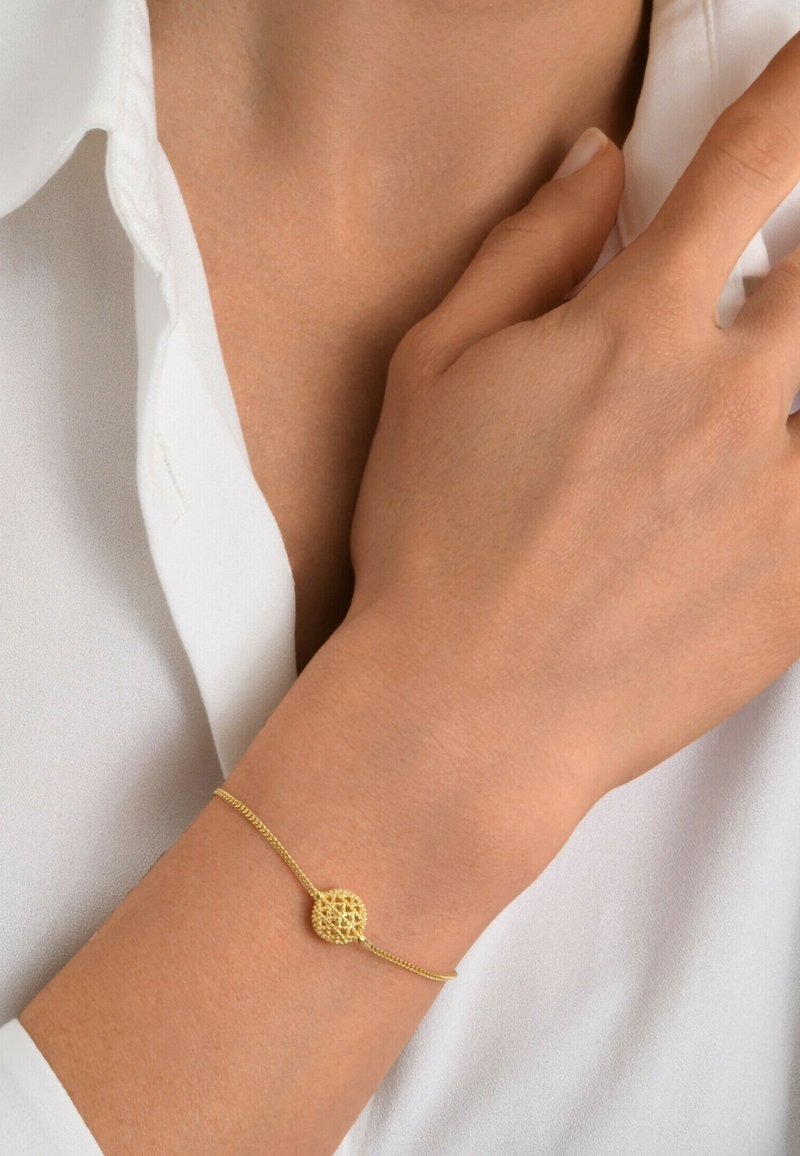 Bracelet en chaîne dorée avec un petit pendentif en filigrane circulaire porté au poignet par-dessus une chemise blanche.