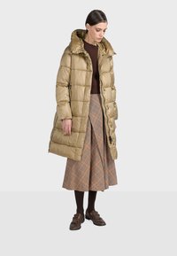 Vadderad beige kappa med huva och knappar framtill, kombinerad med en brun ribbad tröja och en knälång kjol i rutigt mönster i jordnära färger.