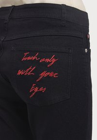 Black denim jeans featuring a back pocket with red embroidered text: "Touch only with your Eyes." Simple hardware accents present.