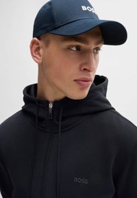 Black zip-up hoodie with a high collar, featuring a subtle logo. Complemented by a navy blue cap with white text. Smooth fabric texture.