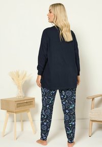 Granatowa bluzka z długim rękawem w połączeniu z czarnymi spodniami z kolorowym wzorem niebieskiego nieba. Miękka tkanina o luźnym kroju. Proste drewniane meble w tle.