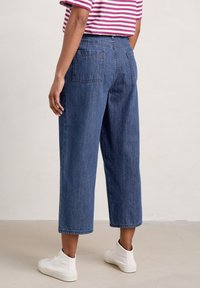 Džínové kalhoty s vysokým pasem a širokýma nohavicema v modré barvě mají dvě zadní kapsy a čistý lem. Kombinované s bílými plátěnými botami.