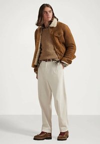 Jeune homme portant une veste en shearling beige, un pull marron, une chemise à carreaux, un pantalon crème et des bottes brunes, se tenant avec une main dans la poche.