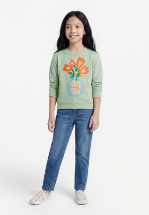 Fille aux longs cheveux noirs portant un sweat-shirt vert avec un motif de bouquet de tulipes, un jean bleu et des chaussures blanches à fleurs sans lacets, debout et souriante.
