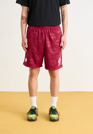 Shorts de sport marron avec une taille élastique, présentant un motif subtil et un détail logo. Portés avec des chaussettes blanches et des baskets colorées.