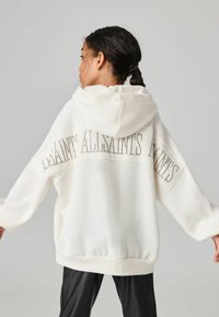 Felpa bianca oversize con cappuccio, caratterizzata da borchie decorative che formano il testo "LA SAINT". I polsini e l'orlo presentano una trama a costine.