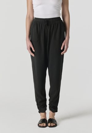 Mujer con pantalones negros sueltos con cintura elástica y sandalias negras de tipo slide, de pie frente a un fondo gris liso.