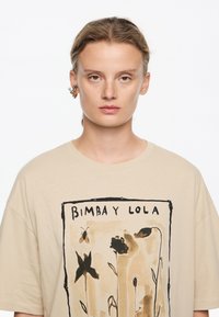 Beež t-särk, millel on suur lilleline illustreerimine maapealsetes toonides. Ülal on tekst "BIMBA Y LOLA" ja peened metallilised aktsendid.