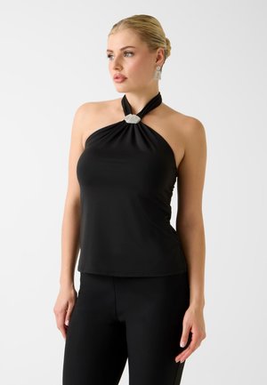 Donna che indossa un top nero senza maniche con chiusura gioiello al collo e pantaloni neri abbinati, posando davanti a uno sfondo bianco.