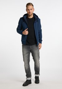 Marineblå zip-up hættetrøje med frontlynlås, sidelommer og justerbare snore, lagdelt over en sort t-shirt og grå jeans. Sorte sko.