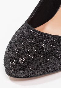 Chaussure plate noire avec un bout arrondi, recouverte de paillettes noires texturées, présentant une doublure intérieure lisse et un bord élastiqué pour le confort.