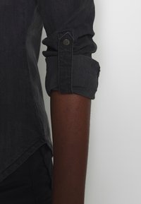 Mörkgrå denimskjorta med uppkavlade ärmar, knäppt manschetter och en lätt asymmetrisk nederkant. Slät textur med subtila vertikala linjer.