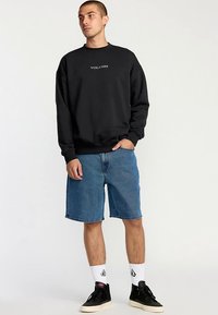 Svart sweatshirt med "VOLCOM"-logotyp, matchad med blåa jeansshorts. Modellen har på sig vita strumpor och svarta sneakers. Enkel design med klassisk passform.