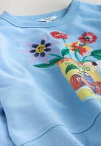 Lichtblauwe sweatshirt met geborduurde kleurrijke bloemen en een geel zakje met het label "Sunbird Peas" aan de voorkant bij de halslijn.