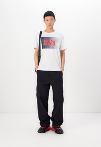 Tricou alb cu mânecă scurtă, cu un imprimeu grafic în roșu și bleumarin, asociat cu pantaloni cargo negri și încălțări negre cu accente roșii.