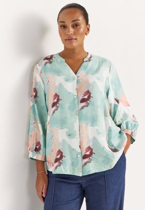 Vrouw draagt een pastelbloemenblouse met knopen en ballonmouwen, gecombineerd met een blauwe spijkerbroek, staand tegen een witte muur, één hand in de zak.