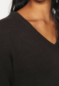 Pull en V marron foncé avec une texture douce et un col côtelé, mettant en valeur une coupe ajustée qui accentue les épaules.