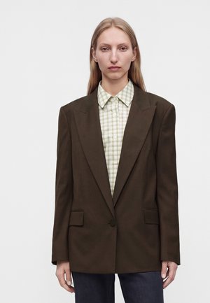 Personne blonde portant un blazer oversize marron foncé sur une chemise à carreaux vert clair boutonnée, debout devant un fond uni.