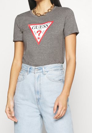 Kobieta w szarej koszulce Guess z czerwonym logo w kształcie trójkąta oraz jasnoniebieskich, wysokich dżinsach, dopełnionych masywnym złotym łańcuszkiem na szyję.