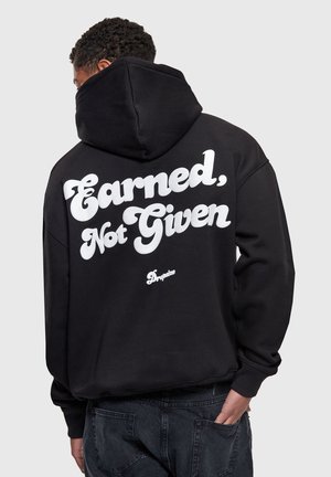 Osoba o kręconych włosach w czarnej bluzie z kapturem z białym napisem „Earned, Not Given” oraz małym logo „Dropsize” na plecach, ręka w kieszeni.