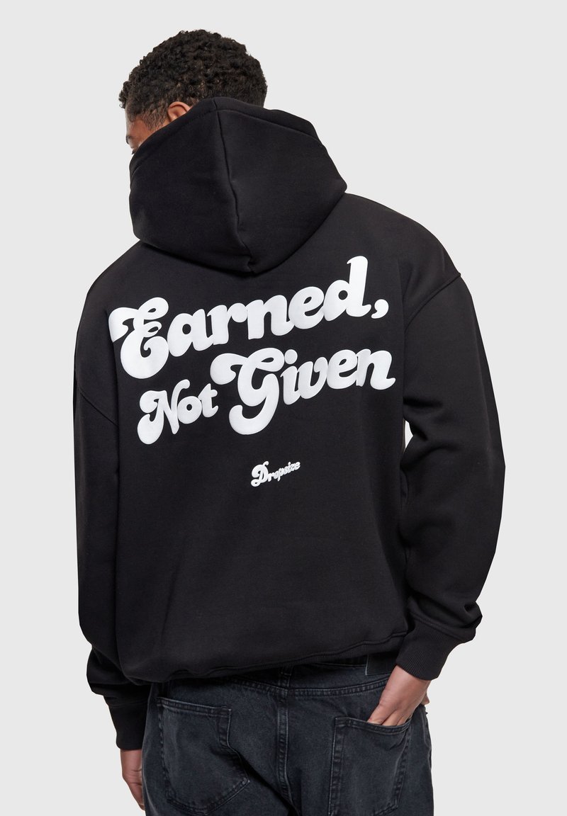 Personne aux cheveux bouclés portant un sweat à capuche noir avec le texte blanc « Earned, Not Given » et un petit logo « Dropsize » au dos, main dans la poche.