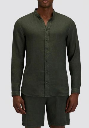 Uomo che indossa una camicia verde scuro a maniche lunghe con bottoni e pantaloncini abbinati contro uno sfondo semplice.