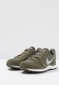Pair of olive green Nike sneakers with white swoosh, suede and mesh details, and white midsoles on a white surface.