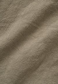 Bézs szövet durva szövött textúrával és enyhe gyűrődésekkel, amelyek finom árnyékokat vetnek a felületre.