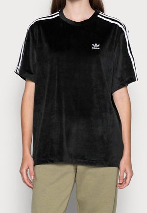 Persona che indossa una maglietta nera in velluto a maniche corte Adidas con strisce bianche sulle spalle e pantaloni beige, in piedi contro uno sfondo neutro.