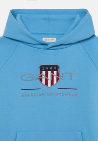 Svetlo modra kapuca z sprednjim žepom, ki ima emblem v rdeči, beli in modri barvi, z vezenim napisom "GANT" in "AMERICAN SPORTSWEAR".