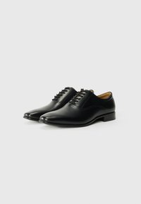 Zapatos de vestir de cuero negro con un acabado suave, punta afilada, seis ojetes y un tacón bajo. Forro interior de color marrón claro visible.