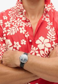 Camisa roja con estampado floral blanco, mangas cortas, cuello abotonado; reloj de pulsera negro con detalles plateados y correa de cuero.
