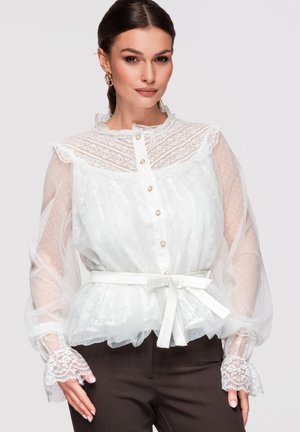 Femme portant un chemisier en dentelle blanche avec des manches longues transparentes et une ceinture à la taille, associé à un pantalon foncé, sur un fond uni.