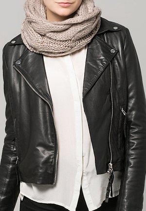 Femme portant une écharpe tricotée beige, une veste en cuir noire et une chemise blanche à boutons sur un fond gris uni.