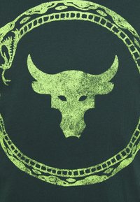 Tela verde con un gran cráneo de toro texturizado en neón amarillo en el interior de un patrón decorativo de serpiente. El diseño es audaz y gráfico.