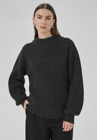Frau mit glattem braunem Haar, die silberne Creolen, einen dunkelgrauen, gerippten Strickpullover und schwarze Hosen trägt und mit einer Hand in der Tasche steht.