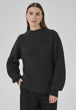 Frau mit glattem braunem Haar, die silberne Creolen, einen dunkelgrauen, gerippten Strickpullover und schwarze Hosen trägt und mit einer Hand in der Tasche steht.