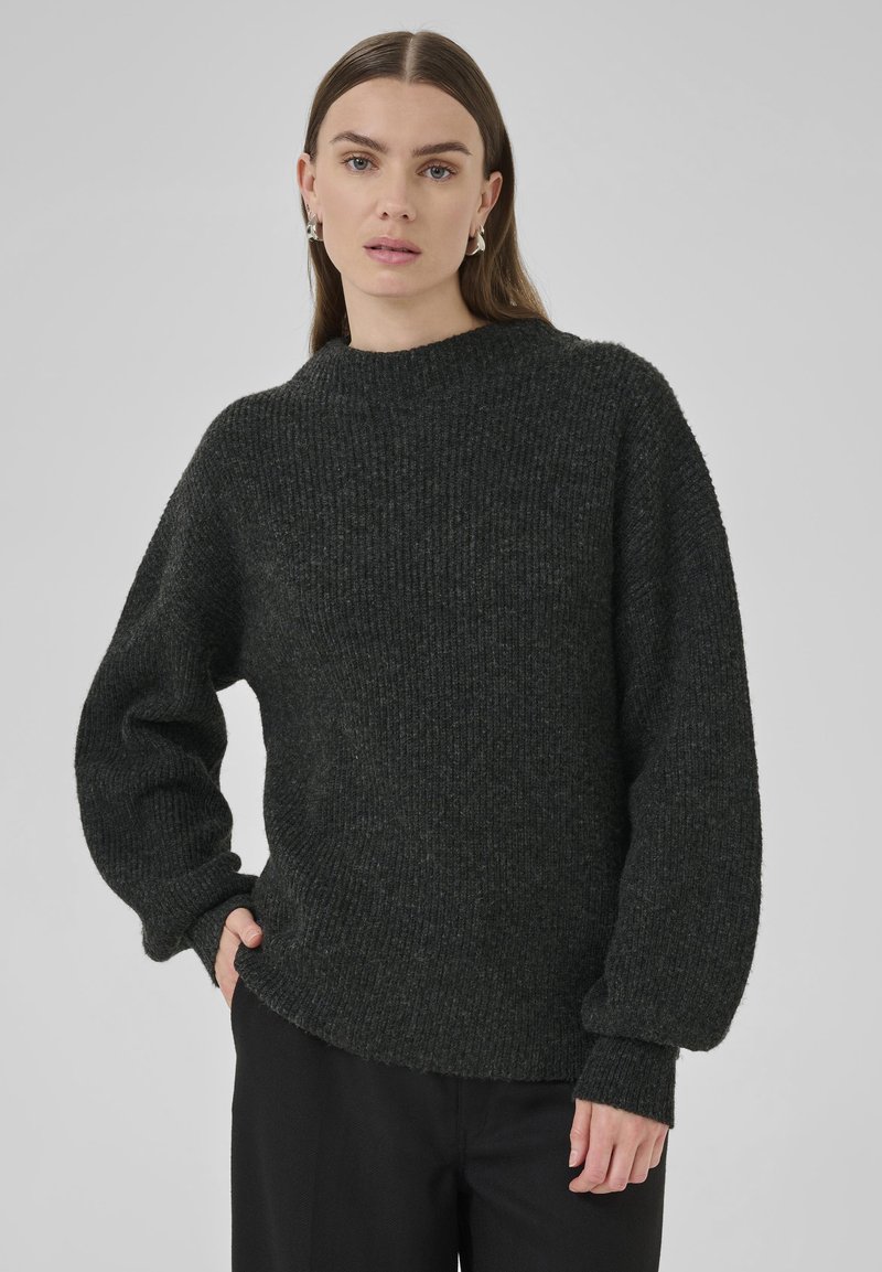 Frau mit glattem braunem Haar, die silberne Creolen, einen dunkelgrauen, gerippten Strickpullover und schwarze Hosen trägt und mit einer Hand in der Tasche steht.